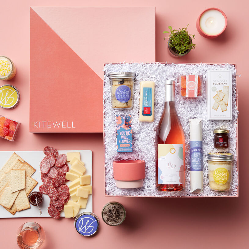 This wine gift box includes Wicked Good Cupcakes Lemon and Cookies & Cream Cupcake Jars, Paddywax Sparkling Grapefruit Candle, and a Modern Sprout Seed Bomb, Dry Salami, Mission Jack Blend, Strawberry Fig Jam, and Sea Salt Flatbread, Sour Peach Hearts, and Rosé.