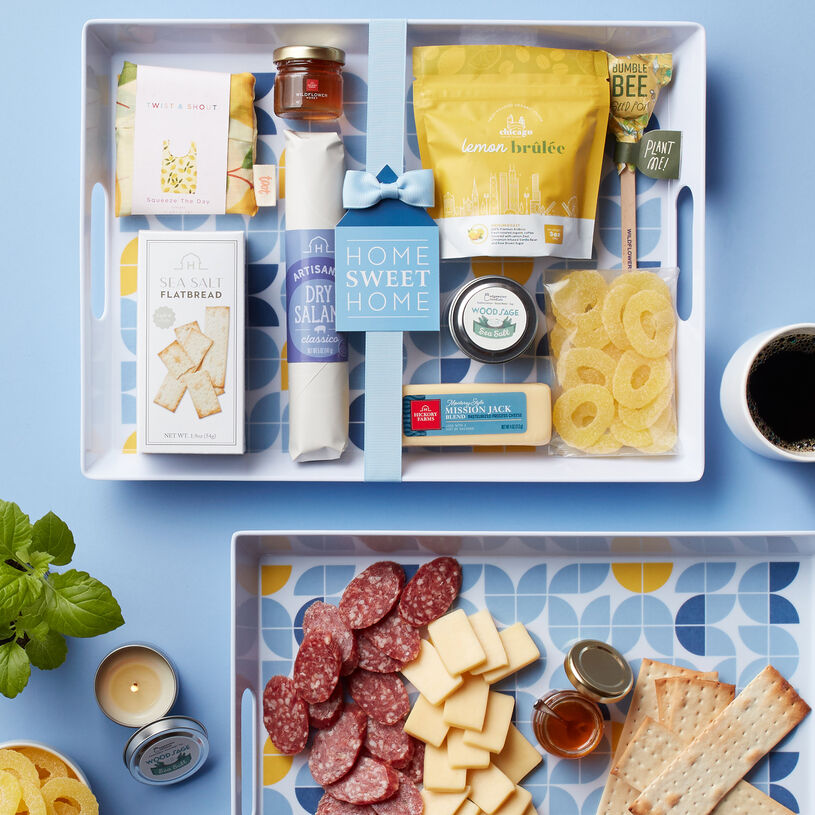 This housewarming gift includes Dry Salami, cheese, crackers, honey, Pineapple Rings, coffee, a Lemon Print Tote, candle, a reusable melamine tray, and a Seed Pop.