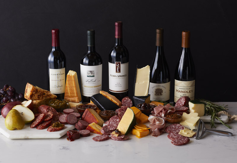 A charcuterie spread of cured meats, cheeses, and fruits in front of a variety of red wine bottles 