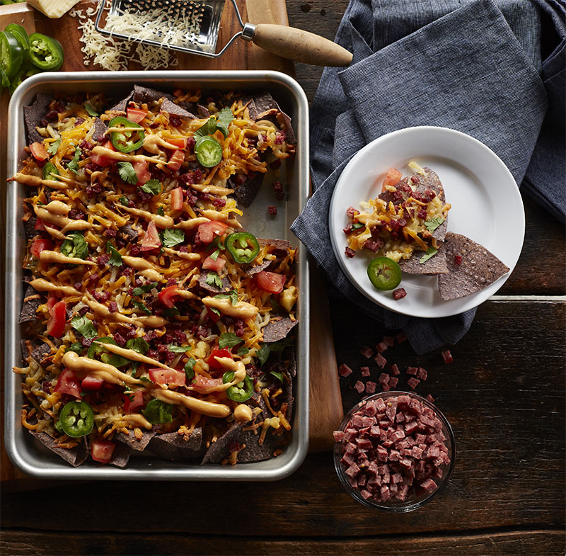 Game day queso with blue corn chips, melted cheese, summer sausage, tomatoes, jalapenos, and chipotle ranch drizzle