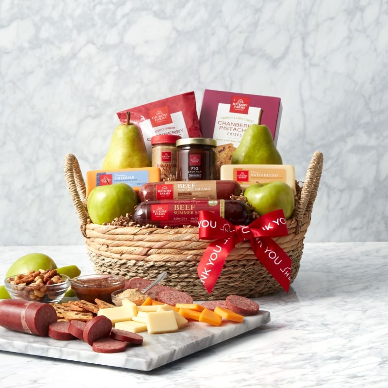 A photo of a gift basket with a thank you ribbon, fruits, sausage, and cracker crisps