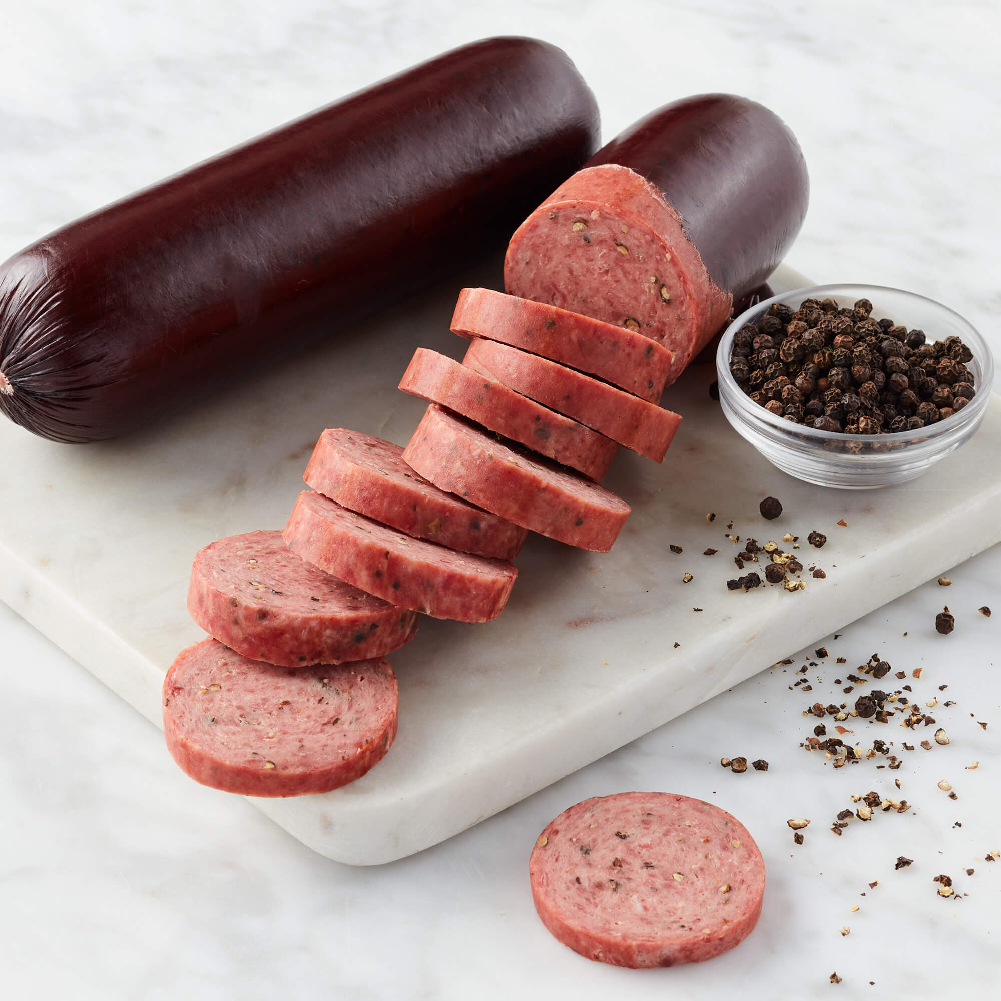 Cracked Pepper Summer Sausage unwrapped and sliced on marble board next to bowl of pepper